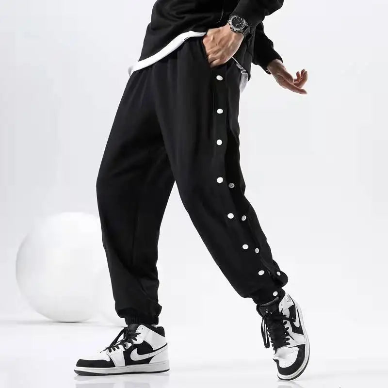 A person wearing black gym pants with white polka dots on the sides, a black sweatshirt, a wristwatch, and black-and-white sneakers poses stylishly against a plain white background.