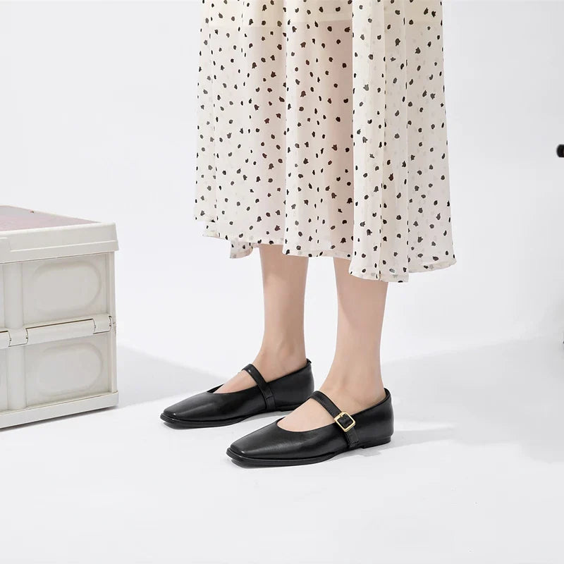 A person wearing a white skirt with a black heart pattern and Augusta Leather Ballet Flats, handmade leather flats with a gold buckle, stands on a white floor near a white textured box.