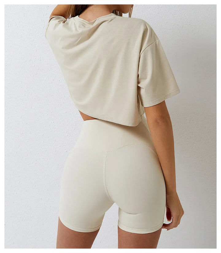 A person stands against a white wall wearing a cotton crop top and high-waisted, fitted beige shorts, with their back facing the camera and one hand resting at their side.