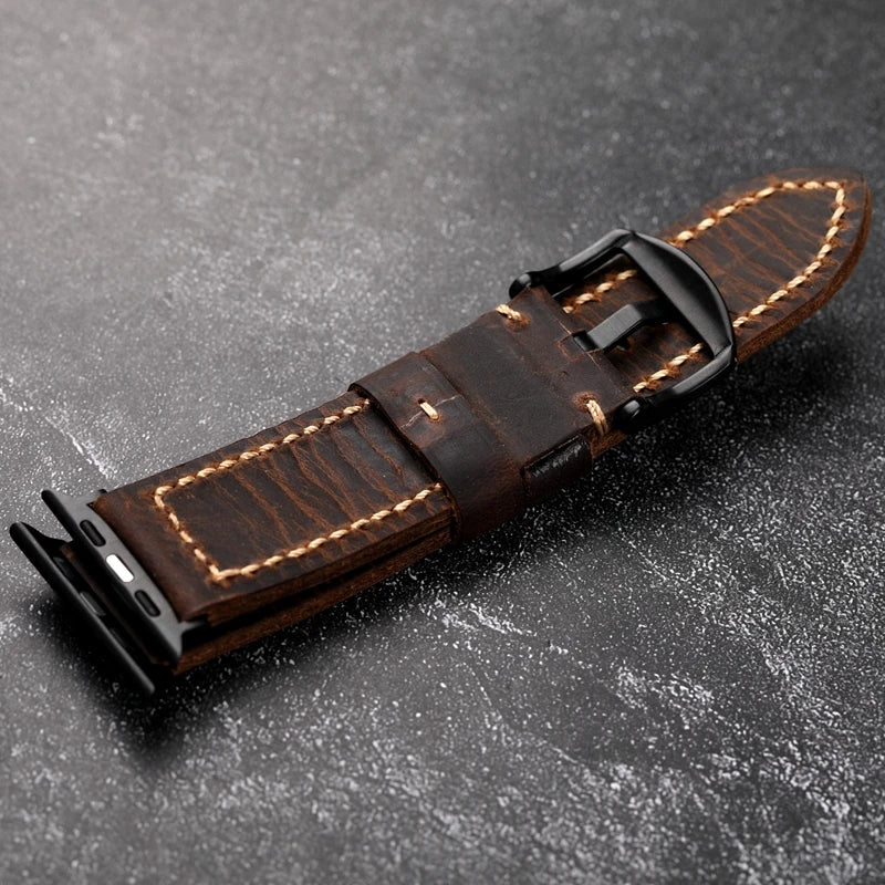 A brown handcrafted Italian cowhide leather watch strap with beige stitching and a black buckle, lying on a dark textured surface. The strap is saddle stitched and designed for a smartwatch.