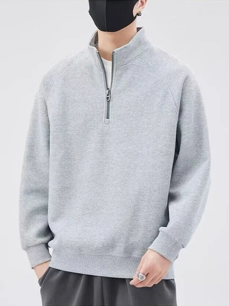 A person wearing a light gray zip-up sweater with a stand-up collar, dark pants, and a black face mask stands against a plain light background. Their hands are relaxed, and the face is mostly covered.