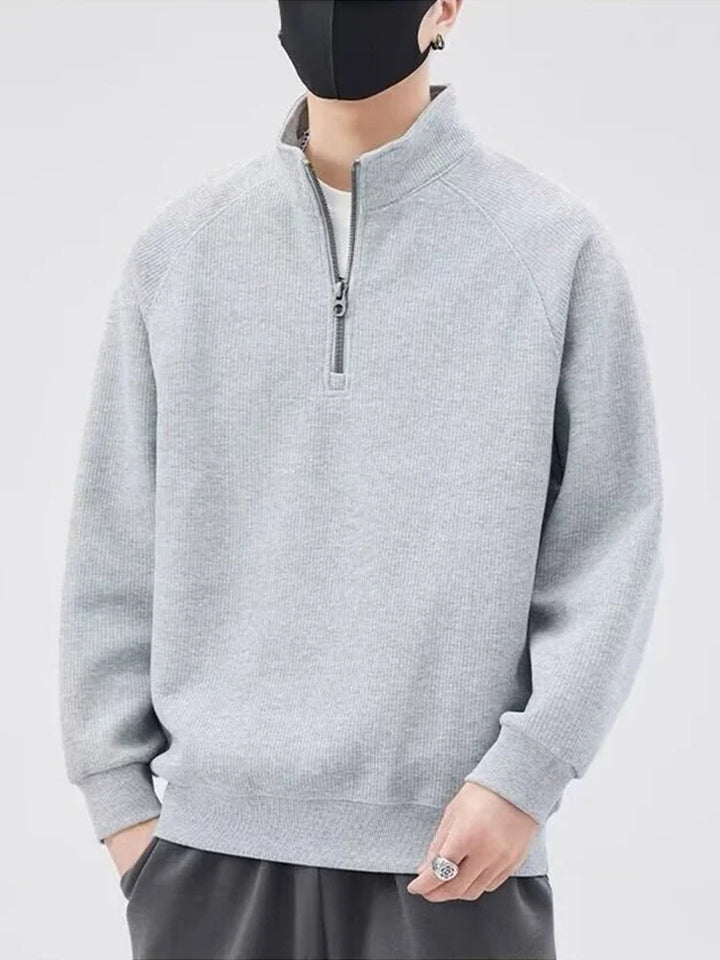 A person wearing a light gray zip-up sweater with a stand-up collar, dark pants, and a black face mask stands against a plain light background. Their hands are relaxed, and the face is mostly covered.