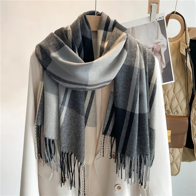 Gray and black plaid scarf in a soft cashmere blend with fringed ends draped over a light-colored coat on a wooden hanger, with a tan quilted vest and a photo in the background.
