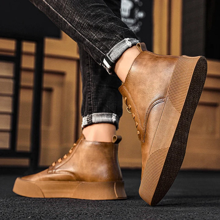 A person wearing cuffed, dark jeans and vintage-inspired high-tops with thick rubber soles and decorative stitching stands indoors on a wood floor. The stylish brown sneakers feature a cushioned footbed for added comfort.
