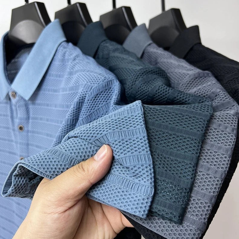 A hand holds the sleeve of a classic polo shirt with textured patterns, while four similar men’s silk polo shirts in blue, teal, and black hang on black hangers in the background.