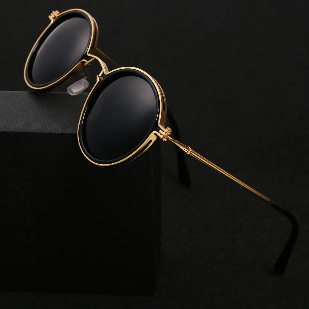 Round Ruby Ray Sunglasses with gold metal frames and black lenses rest on the edge of a black box, set against a dark background. Enjoy stylish UV400 protection wherever you go.