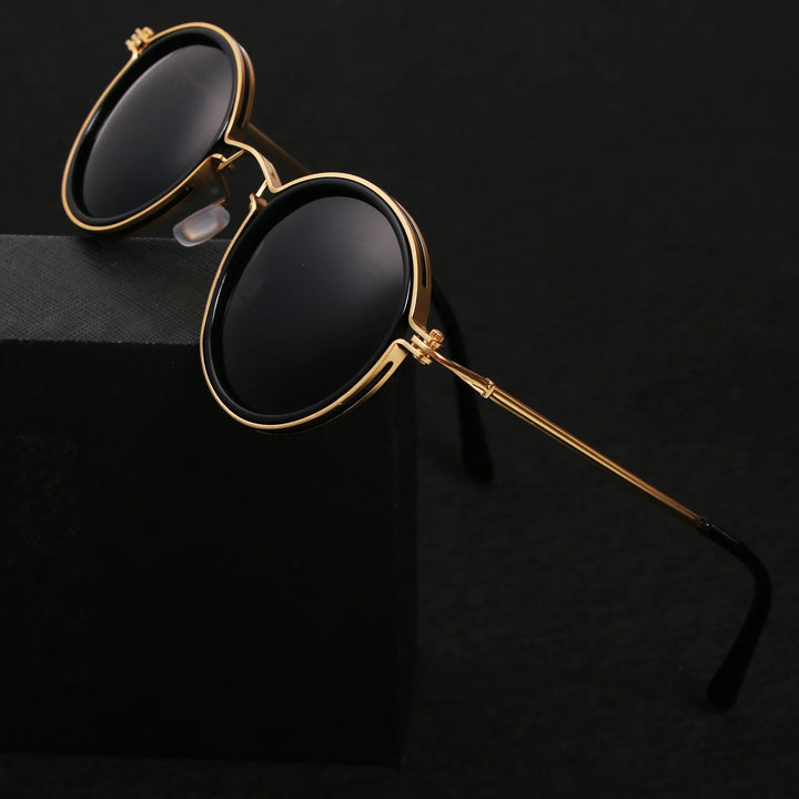 Round Ruby Ray Sunglasses with gold metal frames and black lenses rest on the edge of a black box, set against a dark background. Enjoy stylish UV400 protection wherever you go.
