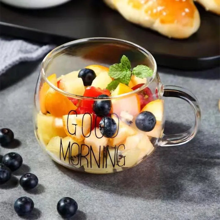 A heat-resistant glass mug with GOOD MORNING printed on it is filled with a colorful fruit salad and garnished with mint; scattered blueberries are nearby, and bread rolls are in the background.
