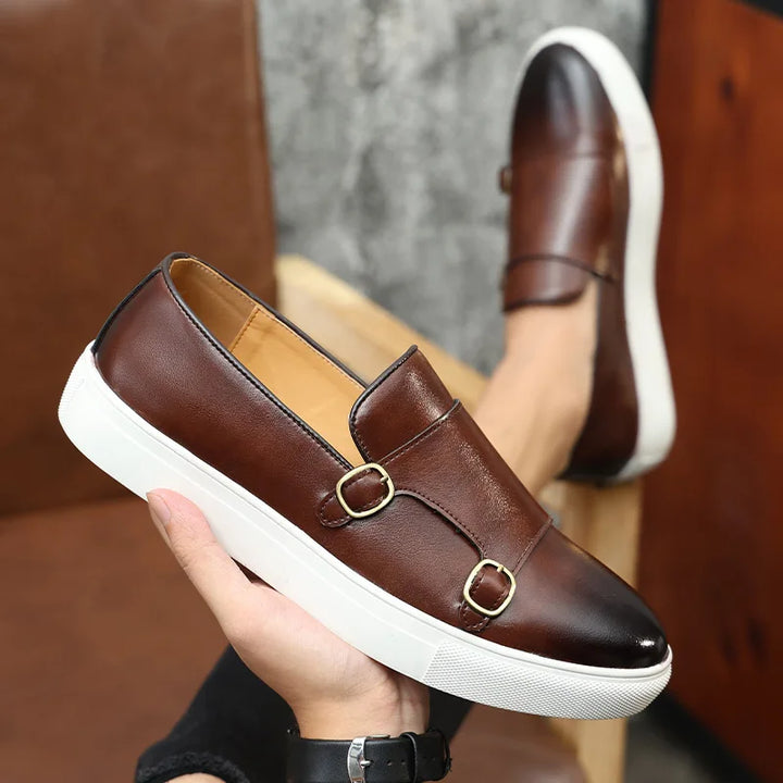 A hand holds a brown leather slip-on loafer with double buckle details and a thick white rubber sole, showcasing classic British fashion. Another matching men’s casual leather shoe is worn on a foot in the background.