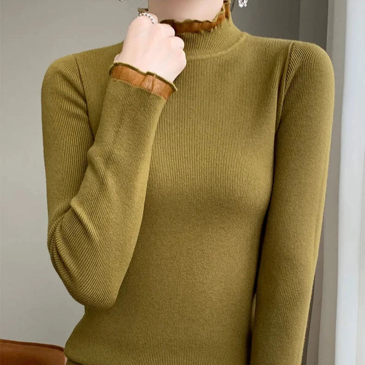 A person wearing an olive green, long-sleeve, ribbed turtleneck sweater with brown ruffled mock neck trim at the collar and cuffs, shown from shoulders to hips, standing indoors.