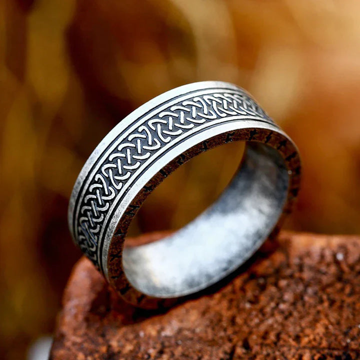 A durable metal ring with intricate Celtic knot patterns engraved around the band, resting on a textured brown surface with a blurred, warm-toned background.