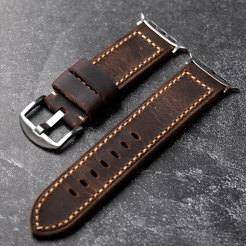 Two pieces of a handcrafted dark brown Italian cowhide leather watch strap, saddle stitched with beige thread and finished with silver metal hardware, are displayed on a textured dark surface.