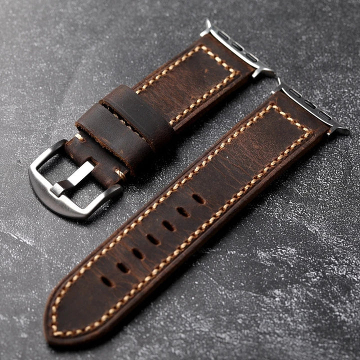 Two pieces of a handcrafted dark brown Italian cowhide leather watch strap, saddle stitched with beige thread and finished with silver metal hardware, are displayed on a textured dark surface.