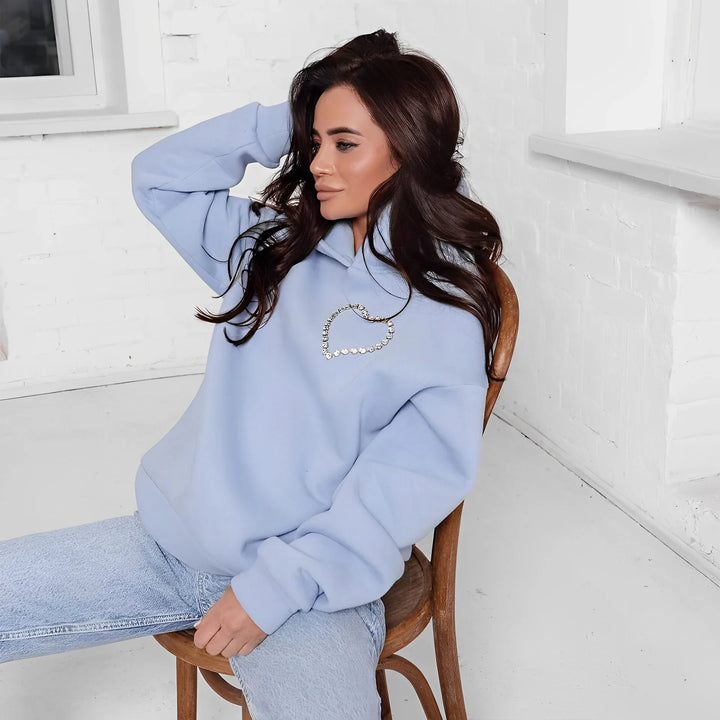 A woman with long brown hair sits on a wooden chair wearing an oversized light blue hoodie with a heart-shaped pearl chain design and rhinestone trim, paired with light blue jeans, against a white brick wall and window background.
