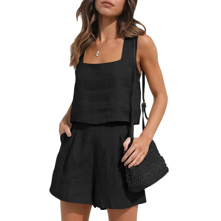 A woman in a contemporary style sleeveless black top and matching high-waisted shorts holds a black textured shoulder bag. She has wavy brown hair and stands with one hand in her pocket against a white background.