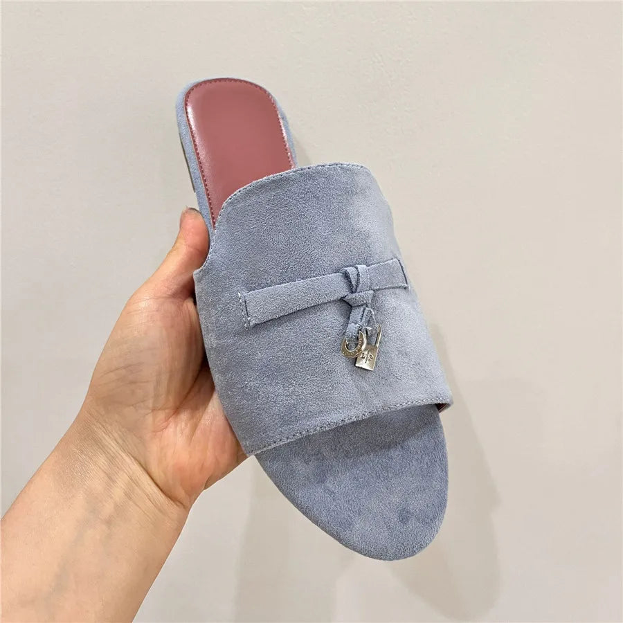 A hand holding a light gray suede slip-on sandal with a cushioned insole and pink lining. The sandal features a decorative strap with a small silver lock charm on the upper for added style.
