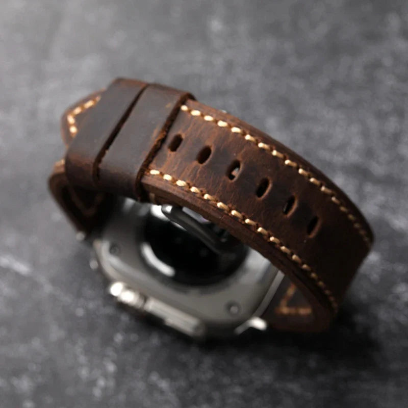 A close-up of a wristwatch with a dark brown, saddle stitched Italian cowhide leather strap, displayed on a dark textured surface. The watch face is mostly out of view, highlighting the craftsmanship of the strap detail.