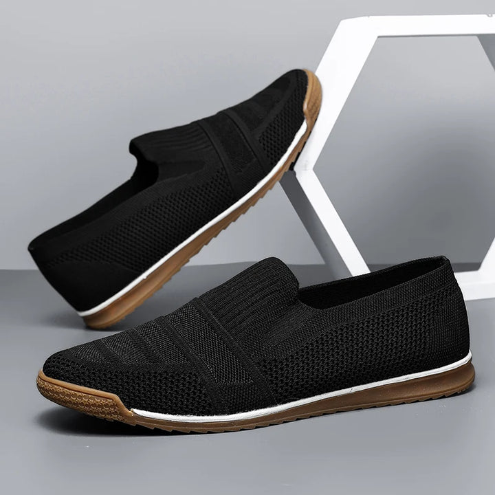 A pair of black air mesh men's casual shoes with brown and white rubber soles, displayed against a gray background with a geometric white prop.