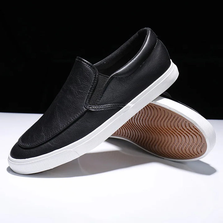 A pair of mens slip-on black sneakers with white rubber-leather soles is displayed, with one shoe standing upright and the other balanced on its heel to showcase the textured sole.