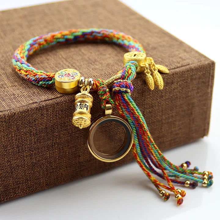 A colorful braided bracelet showcasing Tibetan craftsmanship, with gold charms including a round pendant, a cylindrical charm with inscriptions, and small leaf-shaped charms, displayed on a brown textured box.