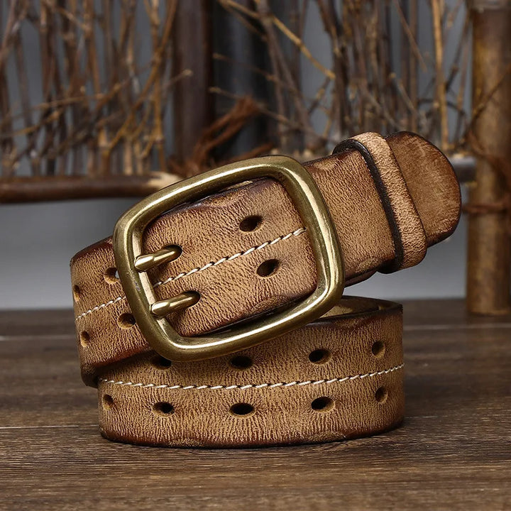 A distressed leather belt with oval-shaped holes and a gold rectangular buckle is coiled on a wooden surface, with rustic wooden sticks and twine in the blurred background.