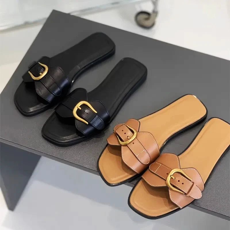 Two pairs of slip-on leather slides with large gold buckles—one in black and the other in tan with a beige insole—are displayed on a gray surface.