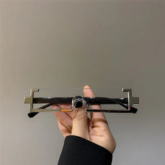 A hand holds a pair of rectangular, frameless eyeglasses with unique metal edges shaped like opposite-facing, geometric zig-zags. These lightweight glasses are set against a plain light gray background.