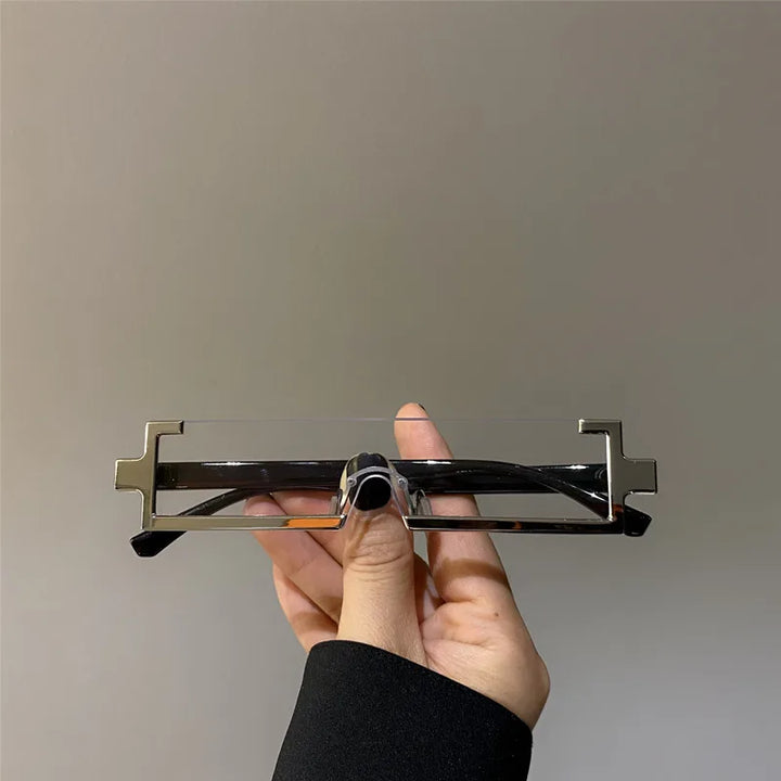 A hand holds a pair of rectangular, frameless eyeglasses with unique metal edges shaped like opposite-facing, geometric zig-zags. These lightweight glasses are set against a plain light gray background.