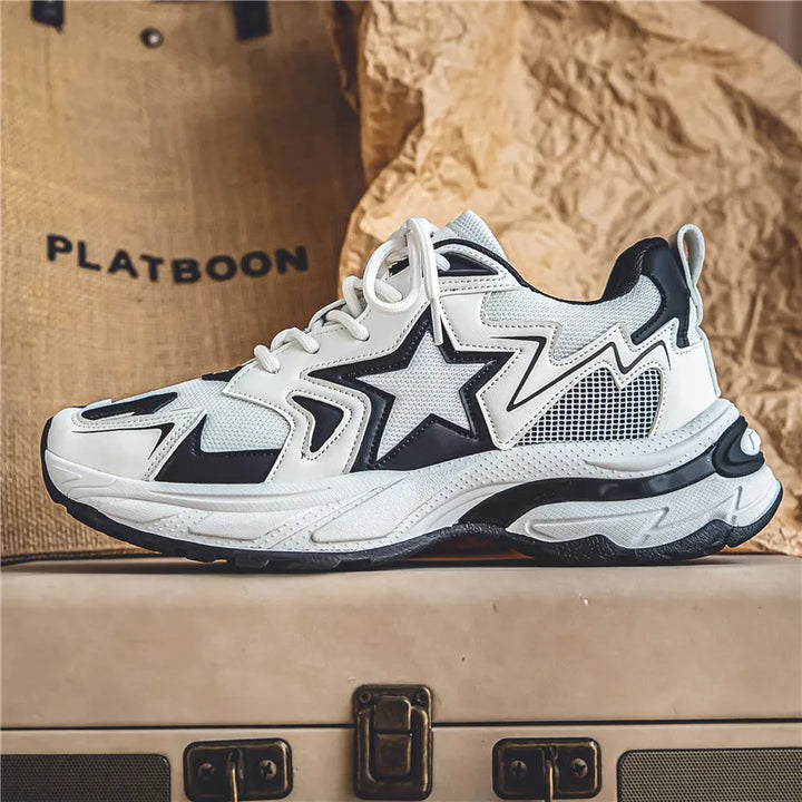 A stylish white sneaker with black accents, a star design on the side, and chunky sole features a durable rubber outsole. Displayed on a suitcase with a beige textured background, the word PLATBOON is partially visible.