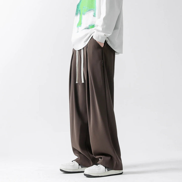 A person wearing a white long-sleeve shirt with a green graphic, loose-fitting brown streetwear joggers with drawstrings, and white sneakers stands against a plain white background.