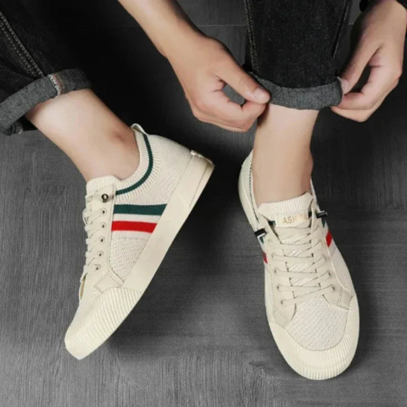 A person rolling up the cuffs of their dark jeans while wearing beige sneakers with green and red stripes and a durable rubber outsole, standing on a dark wooden floor.