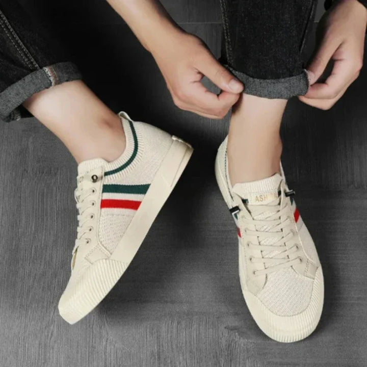 A person rolling up the cuffs of their dark jeans while wearing beige sneakers with green and red stripes and a durable rubber outsole, standing on a dark wooden floor.