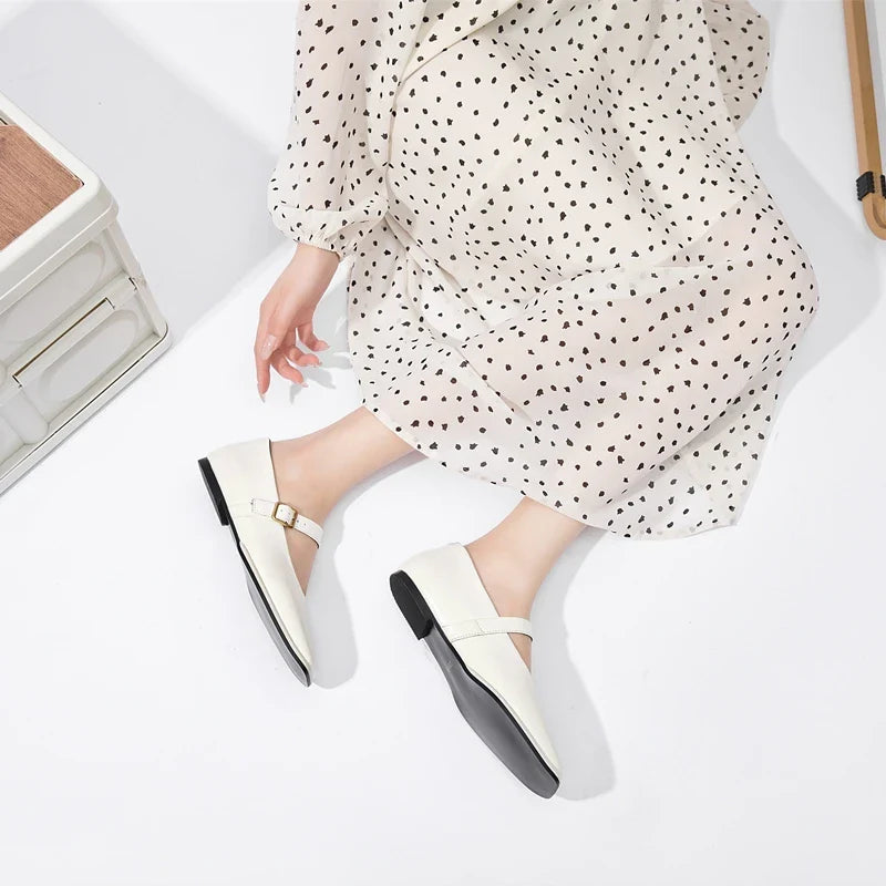 A person wearing a white, long-sleeved, polka-dotted dress is sitting on the floor with legs extended, showing off chic white Mary Jane shoes with a buckle detail.