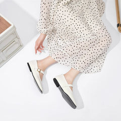 A person wearing a white, long-sleeved, polka-dotted dress is sitting on the floor with legs extended, showing off chic white Mary Jane shoes with a buckle detail.