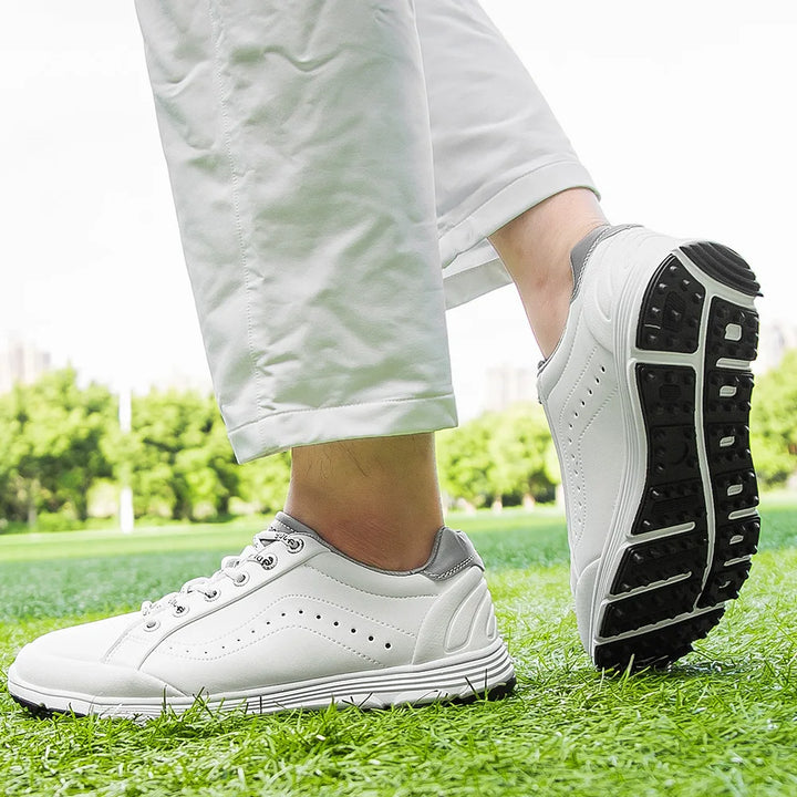 A person wearing white pants and advanced cleat technology golf footwear with black soles stands on green grass outdoors, with trees and a city skyline visible in the blurred background.