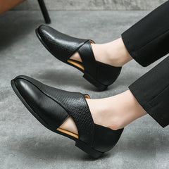 A person wearing low profile loafers with textured side panels, cut-out details, and a durable outsole, paired with black pants, is seated on a gray floor.
