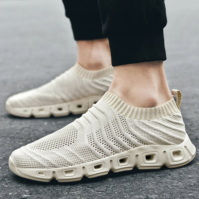 Close-up of a person wearing beige slip-on athletic sneakers with a cushioned sole and mesh upper, standing on a paved surface. The shoes have a modern, breathable design with a sock-like fit, and the person is wearing black pants.