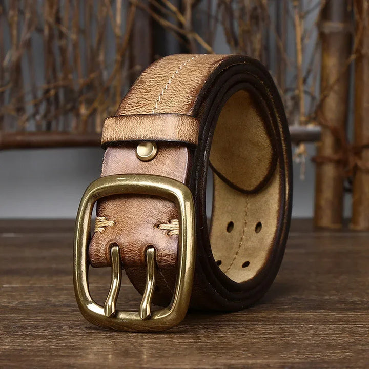 A genuine leather belt with a copper buckle is rolled up and displayed on a wooden surface, set against a blurred background featuring dried branches, reflecting heritage craftsmanship.