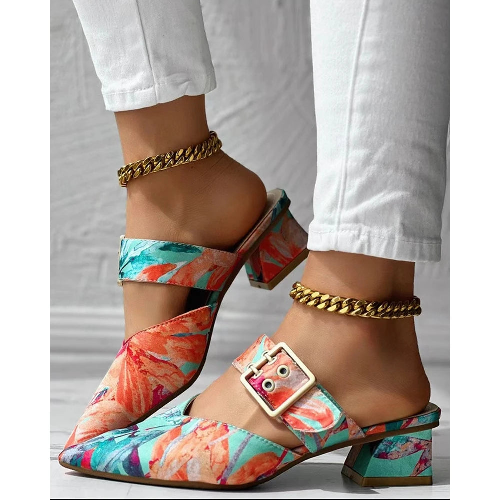A person wearing colorful floral heeled mules slippers with a gold buckle and square heel, paired with white pants and gold chain anklets, standing on a gray floor. Perfect summer shoes for a stylish day out.
