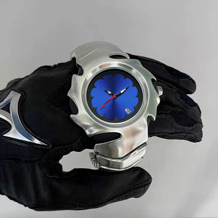 A person wearing a black glove with a silver accent holds a stainless steel, scratch-resistant watch featuring a blue, scalloped-edged face, red hands, and a small date display on a plain gray background.