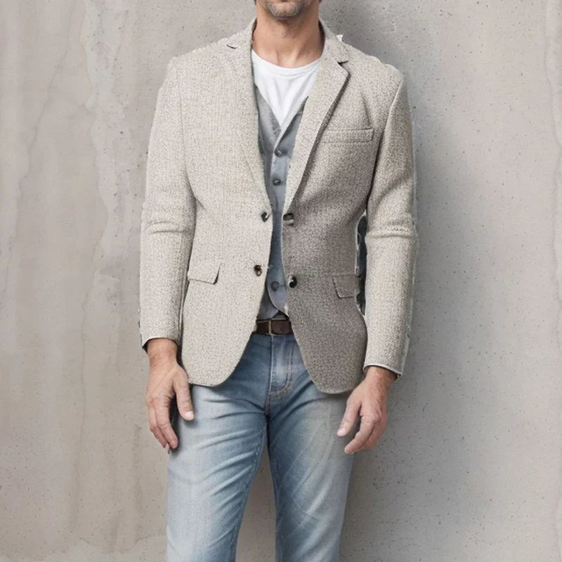 A man stands against a light gray textured wall, wearing a light beige blazer over a gray vest and white shirt, paired with tapered fit light blue jeans. His face is partially out of frame.