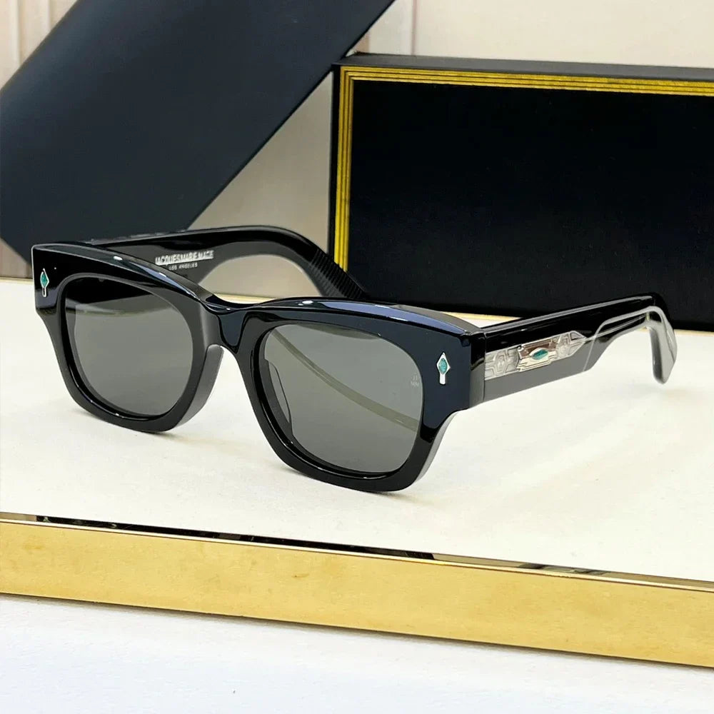 A pair of black rectangular retro sunglasses with dark lenses displayed on a white surface. The temples feature silver and turquoise accents, and there is a gold-trimmed black box in the background.