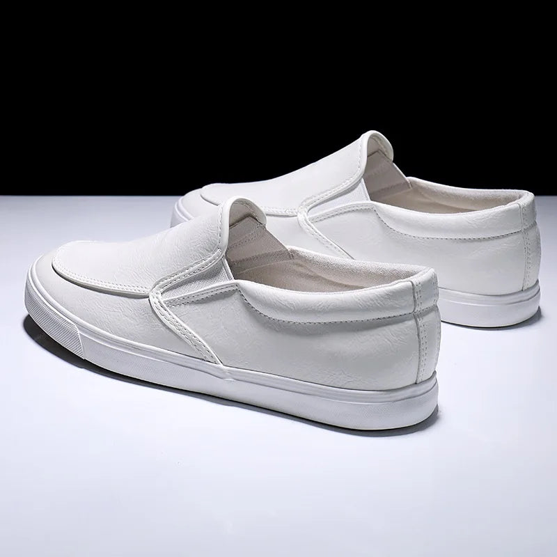A pair of mens slip-on white sneakers with rubber-leather soles, displayed on a glossy white surface against a black background. The shoes are positioned at slight angles, showcasing both the side and back views.
