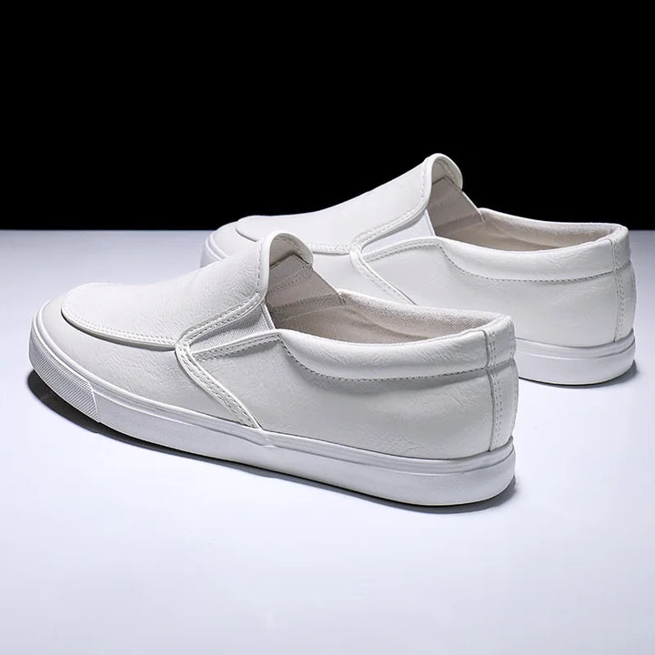 A pair of mens slip-on white sneakers with rubber-leather soles, displayed on a glossy white surface against a black background. The shoes are positioned at slight angles, showcasing both the side and back views.