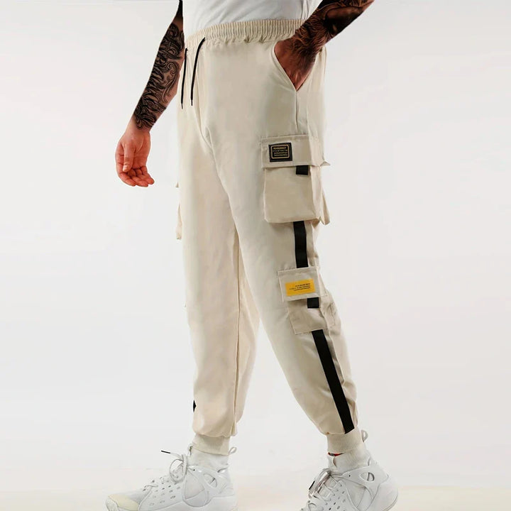 A person wearing light beige cargo jogger pants with an elastic waist, ribbed ankle cuffs, side pockets, black stripes, and yellow patches, paired with white sneakers and socks, stands against a plain white background.