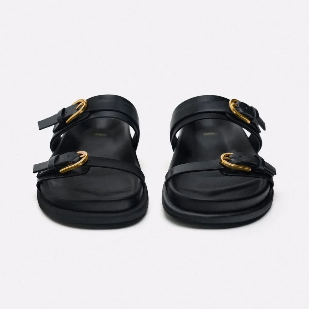 A pair of black leather sandals with a leather upper, wide straps, and gold buckles, viewed from the front on a plain light background.