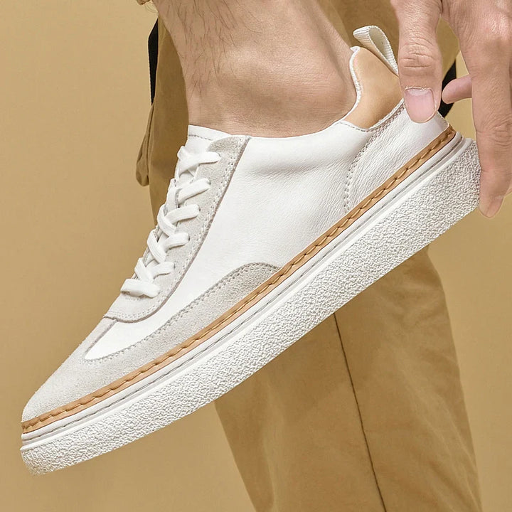 A hand holds a white sneaker with beige accents, tan trim detail, and a textured white sole. Crafted as genuine leather shoes, they feature a cushioned insole for added comfort. The person wears tan pants against a solid beige background.