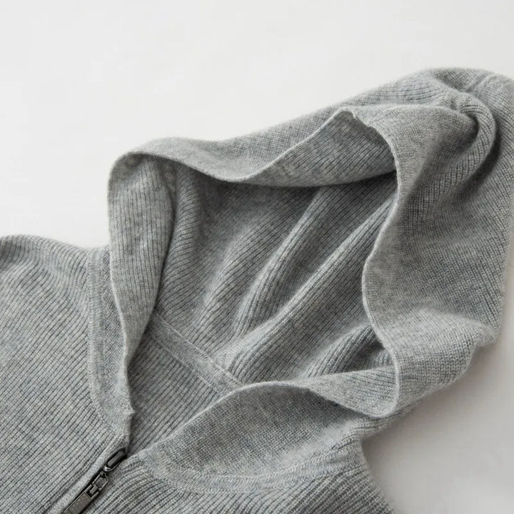 A close-up of a light gray, ribbed knit cashmere hoodie with a zipper, showing the luxurious cashmere texture and lightweight insulating fabric, with the hood laid flat on a white surface.