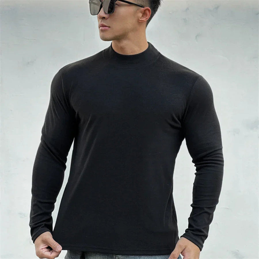 A man wearing a close-fitting, long-sleeve black shirt with a mock neck collar and sunglasses stands against a light gray, textured background. He looks slightly to the side with his hands by his sides.
