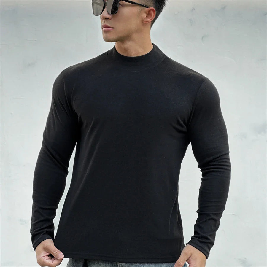A man wearing sunglasses and a fitted, long-sleeve black shirt with a mock neck collar stands against a light gray background.